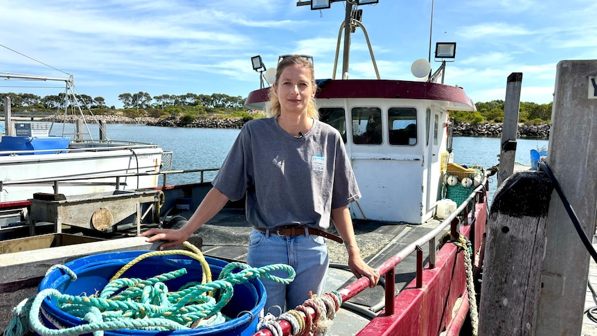 A serious young blonde woman, hair tied, glasses on top, grey tee, jeans, stands on a fishing boat. Blue sky, scattered clouds.