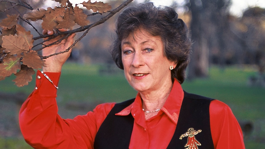 Joy McKean stands beneath a tree in a part, holding onto a branch. She wears a red top and black vest.