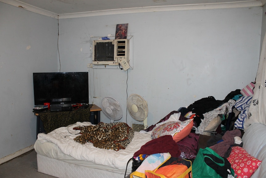 A bed is piled with clothing. Two fans sit next to it along with a TV. There are cracks in the paint and cobwebs on the walls.