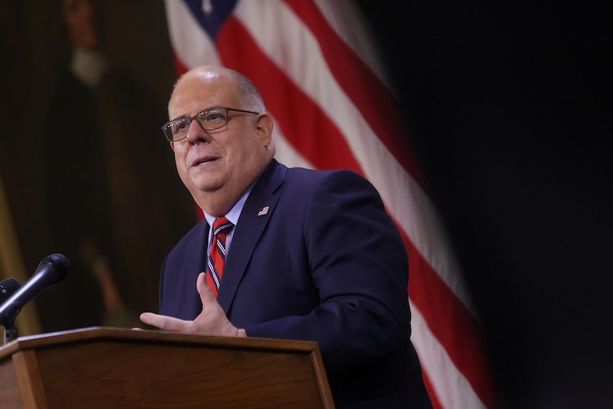 A bald, heavy-set middle-aged white man in a suit speaks behind a lectern in front of an American flag.