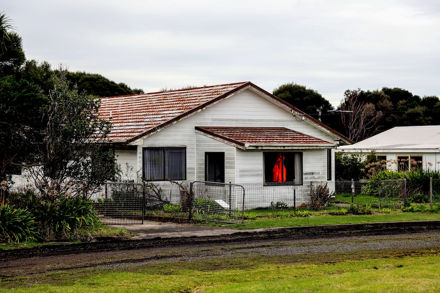 A white timber house with a high vis jacket in the window and grass either side of a dirt road