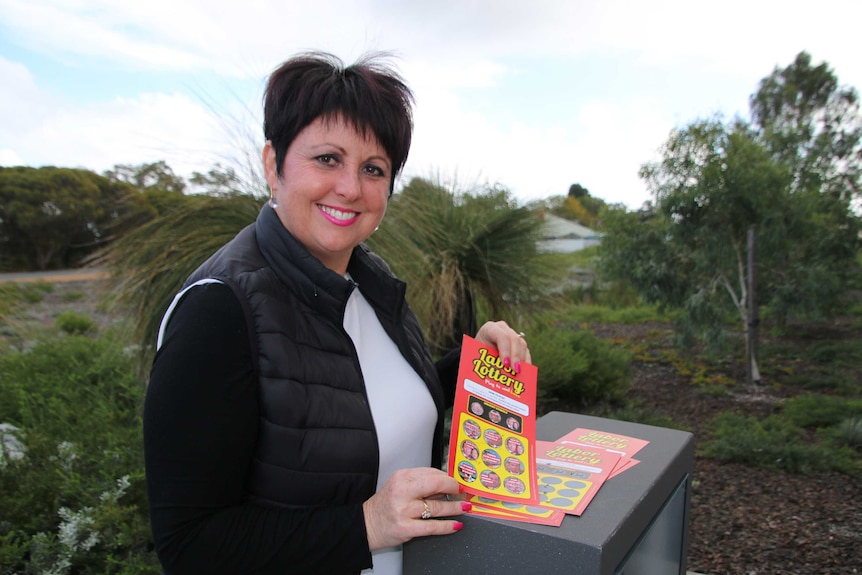 Liberal Darling Range by-election candidate Alyssa Hayden stands smiling near bushes holding a red 'Labor Lottery' scratchy.
