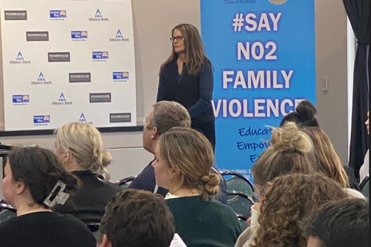 A woman stands in front of a say no 2 family violence banner speaking to high school teachers