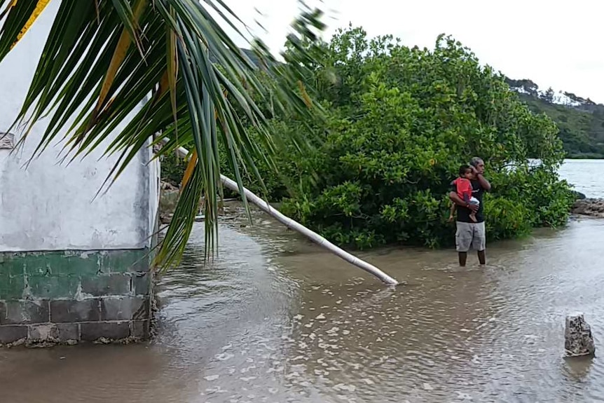 A man stands with his ankles in water and holds his grandchild as sea water inundates his house.