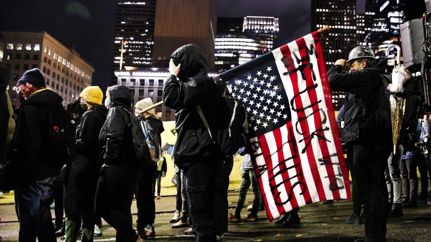 A demonstrator wearing a hoodie carries an American flag with the words 'fuck this' painted on it