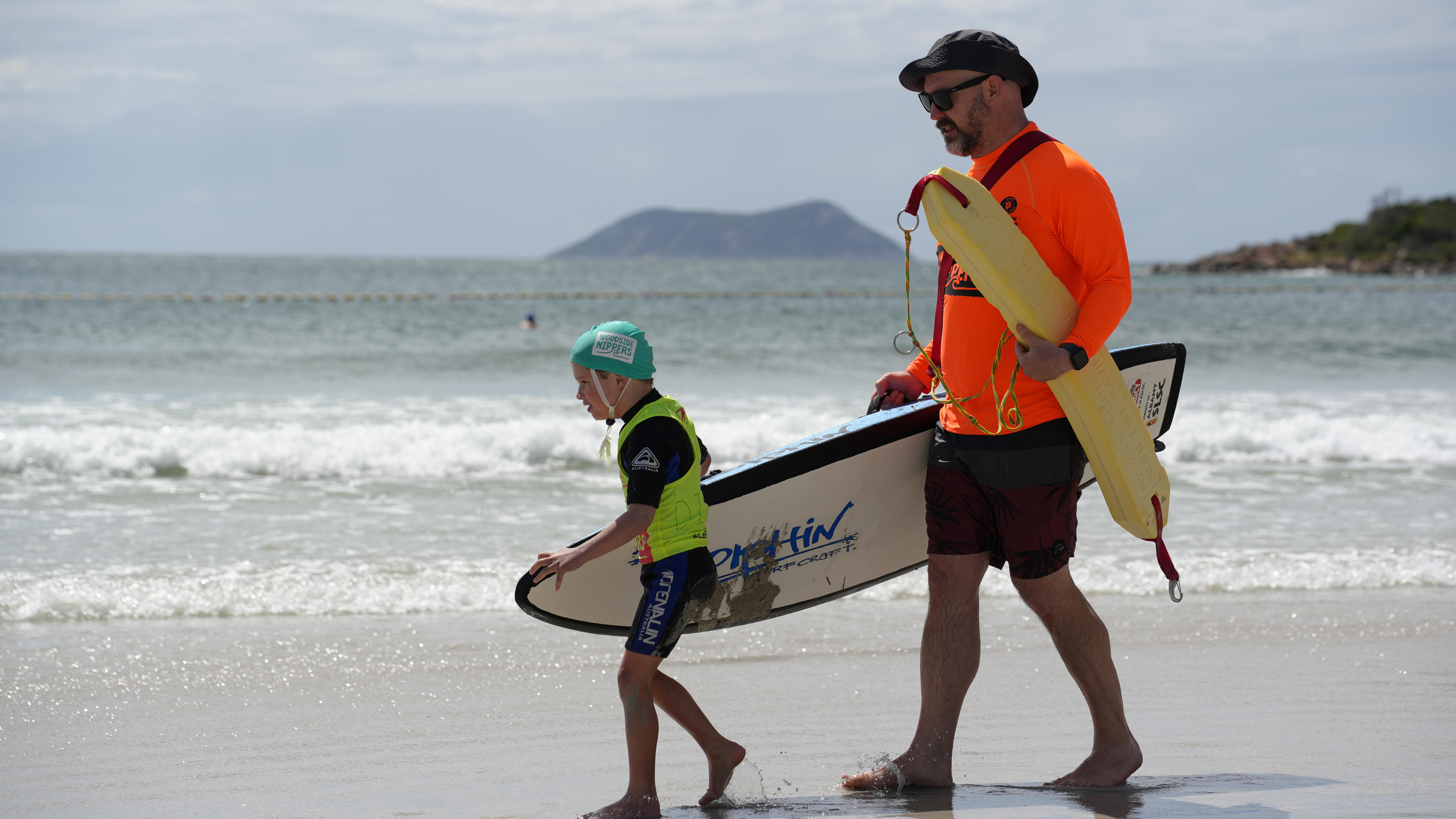 Un niño y un hombre adulto caminan por la costa con una tabla de surf