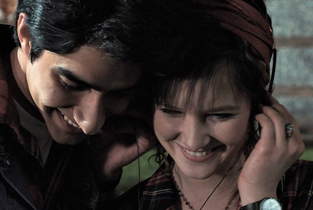 Viveik Kalra and Nell Williams smile downwards while standing close together to listen to music from one pair of headphones.