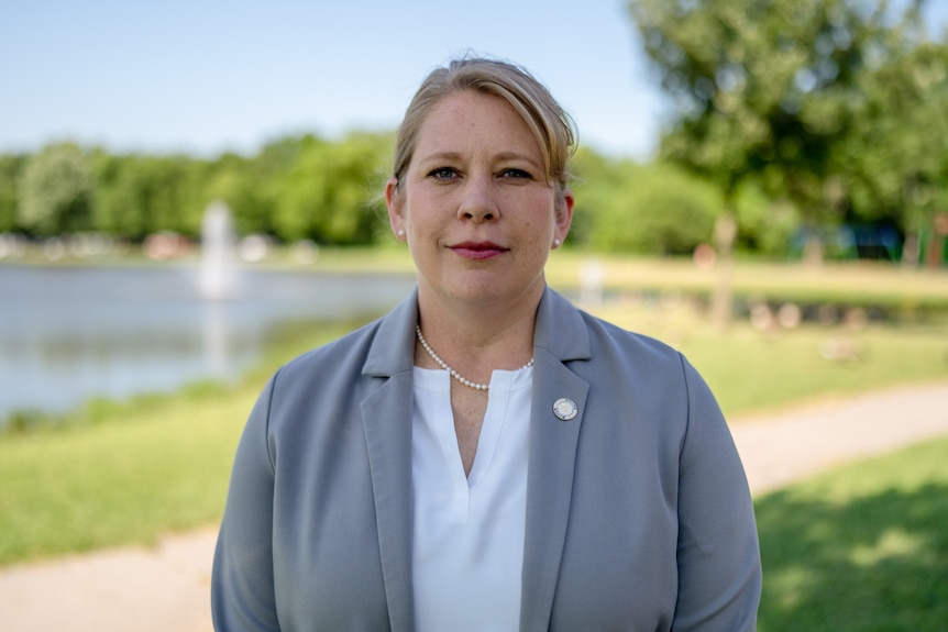 A woman in a grey blazer, white shirt and pearl necklace stands outside in a park next to a lake