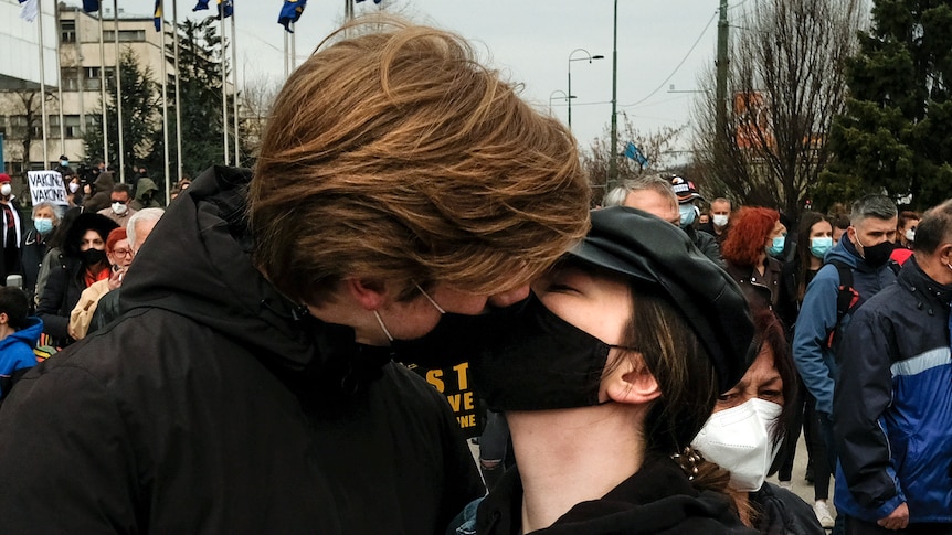 A young woman and a young man, both wearing cloth face masks, kiss in front of a large crowd.