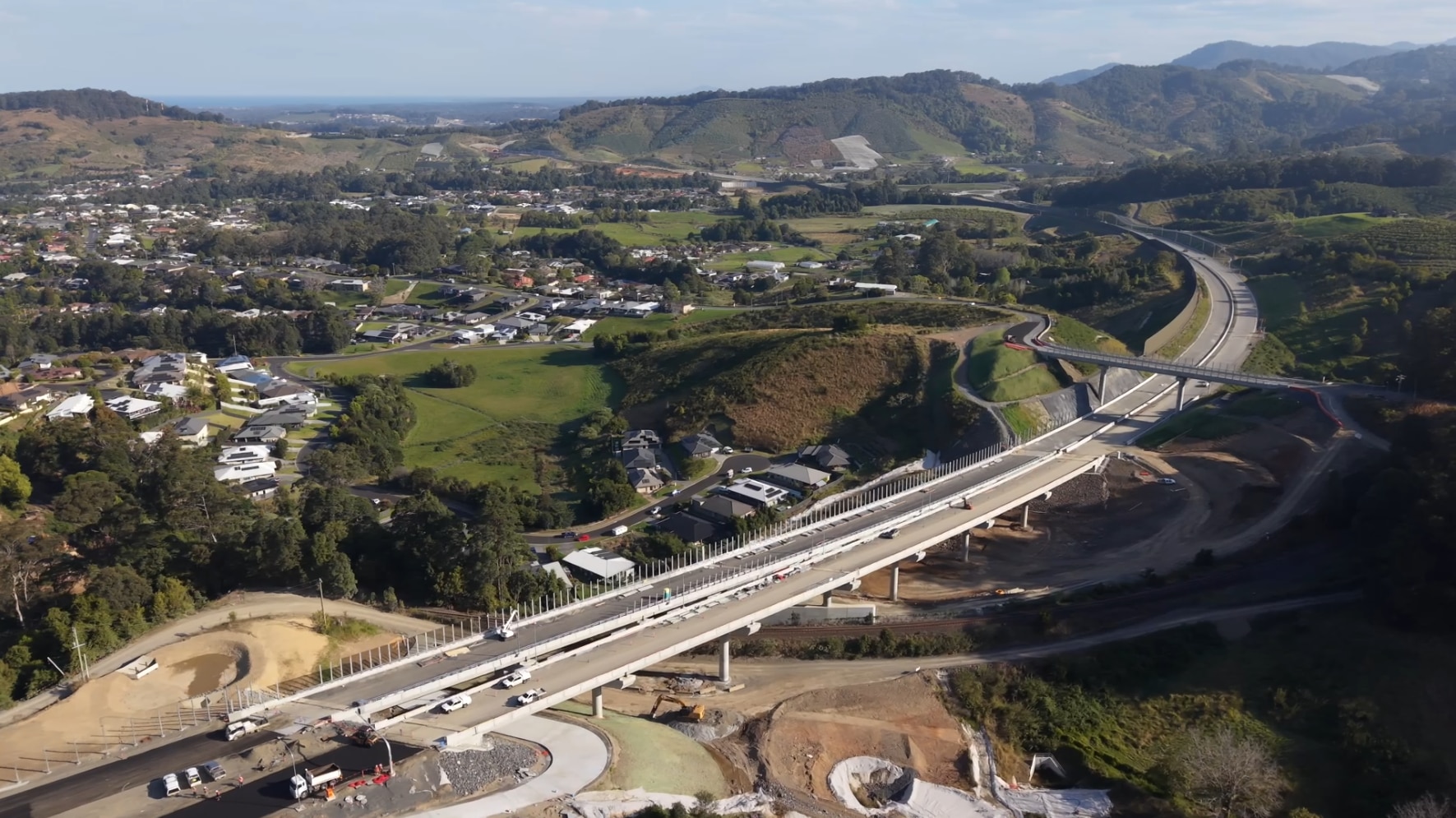Aerial view of road construction stretching into hills and around coastal settlement.