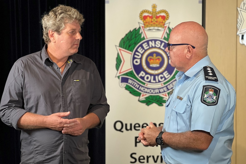 A man wearing a grey shirt speaks with a male police officer in front of a Queensland Police banner.