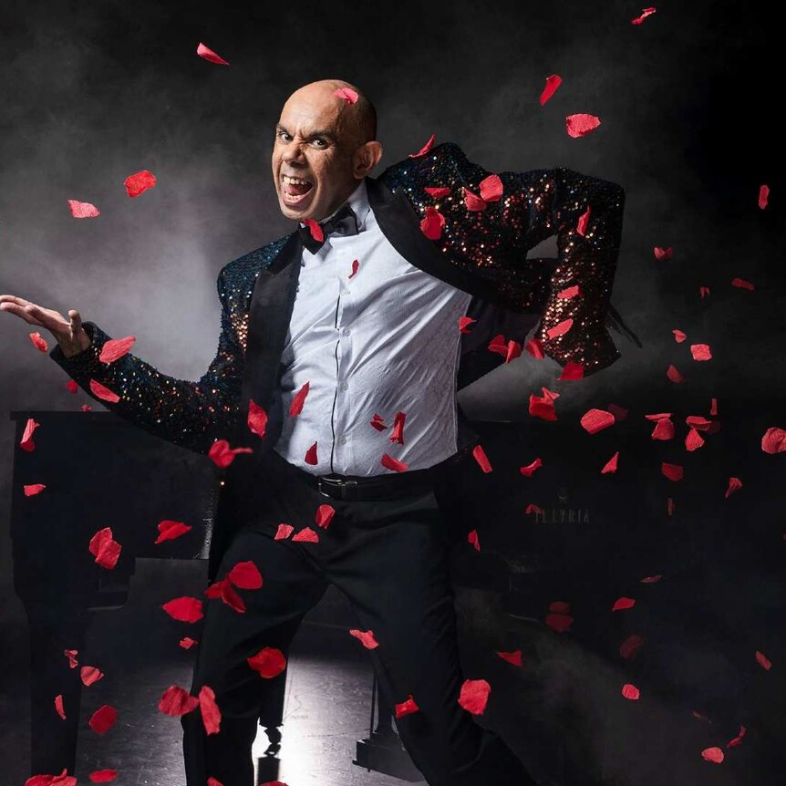 Steven Oliver is a First Nations actor, comedian, singer and performer. He is stood in a sequin tuxedo with a black background.