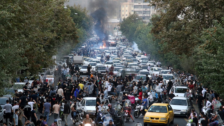 A street is blocked by protesters as smoke and flames rise in the background.