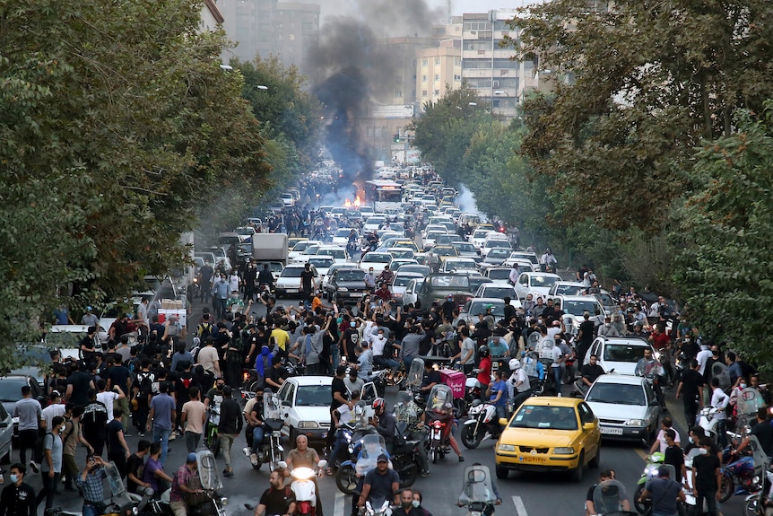 A street is blocked by protesters as smoke and flames rise in the background.