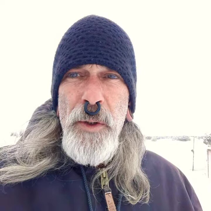 A man with a septum piercing and white beard and long hair wearing a beanie outside while it snows