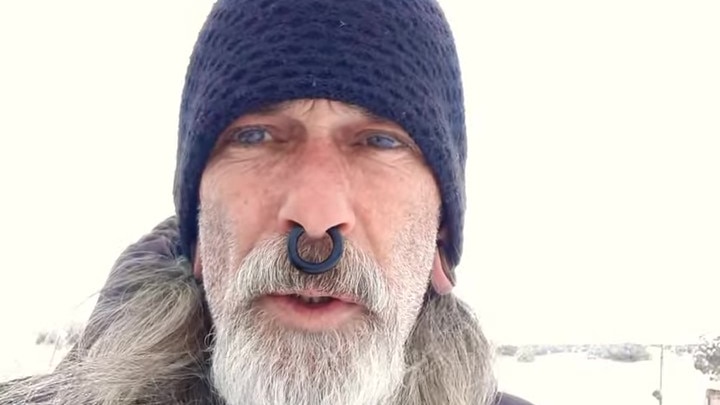 A man with a septum piercing and white beard and long hair wearing a beanie outside while it snows