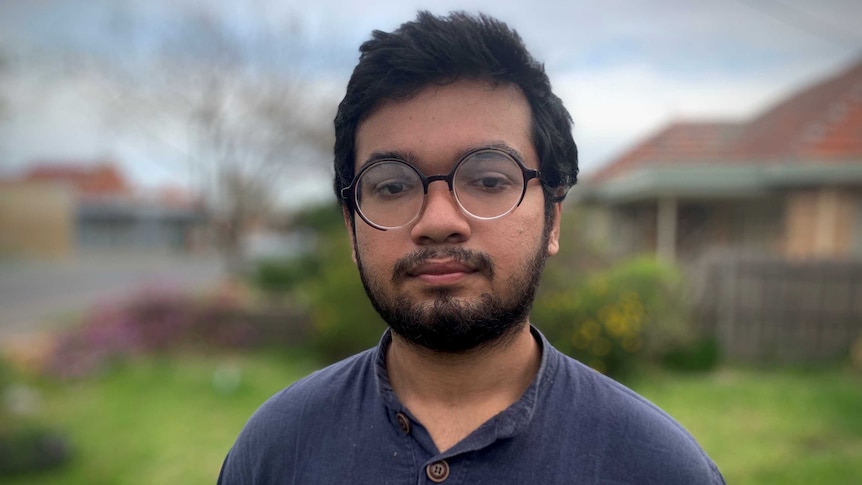 A young man with glasses and a short beard wears a blue top and looks into the camera