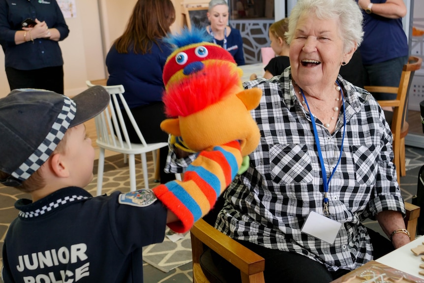An image of Marcia, 85, with short white hair, smiling at a boy with a colourful puppet on his right arm raised  towards her