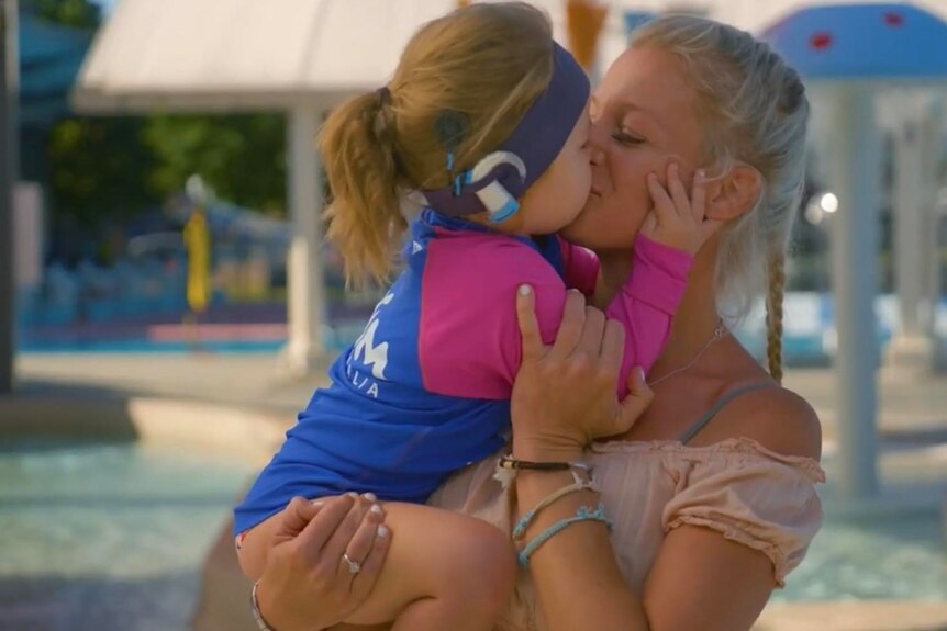 A young girl with a cochlear implant kisses her mothers face. They are standing in a water park. 