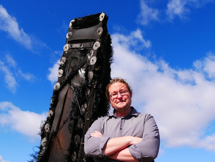 a man stands next to space junk with his arms crossed