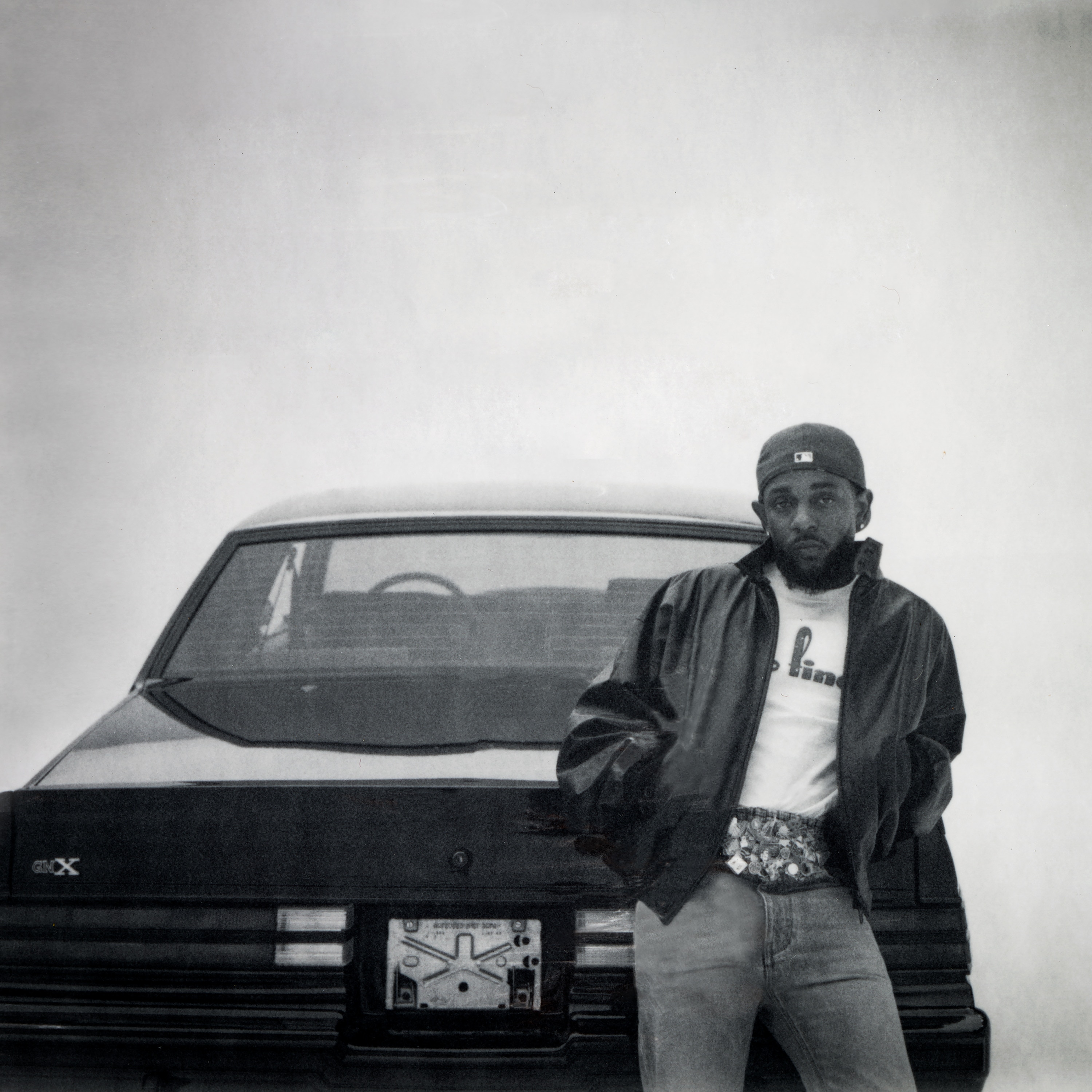 Black and white photo of rapper Kendrick Lamar in street wear leaning on the boot of a 1987 Buick GNX car