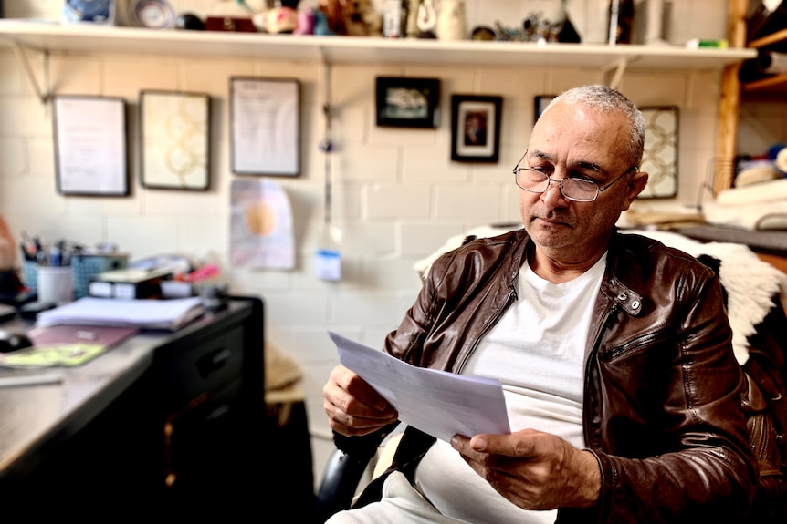 A man wearing a leather jacket and glasses looks at paperwork.