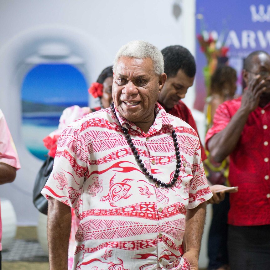 Bob Loughman walks through a room wearing a red and white Pacific shirt and a beaded necklace.
