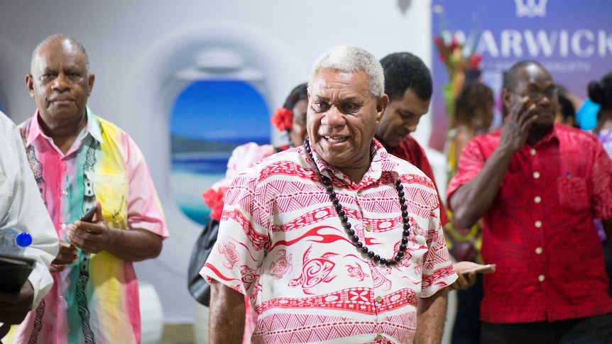 Bob Loughman walks through a room wearing a red and white Pacific shirt and a beaded necklace.