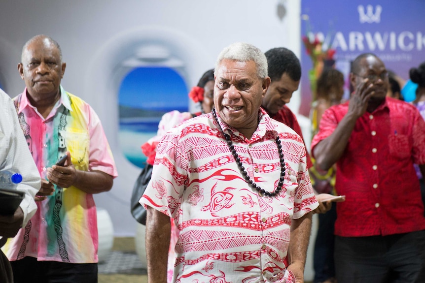 Bob Loughman walks through a room wearing a red and white Pacific shirt and a beaded necklace.