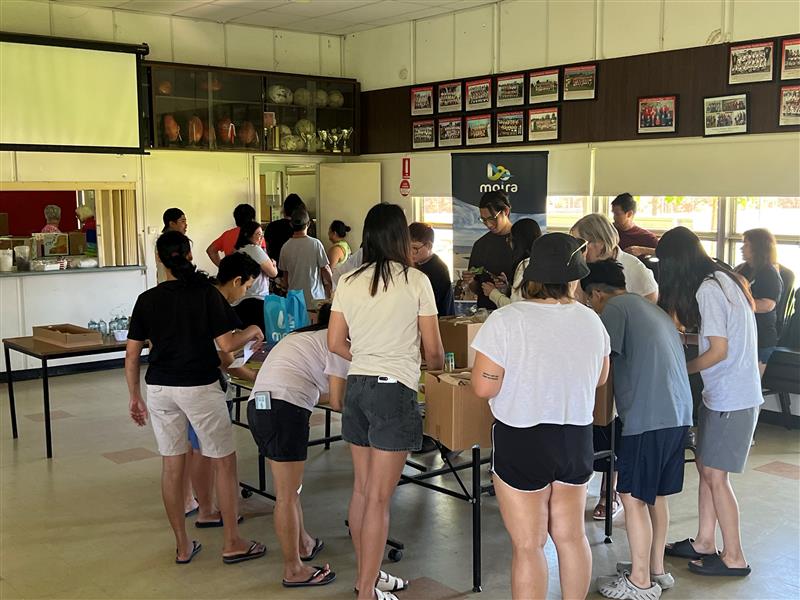 A group of people stands around a table covered with cardboard boxes in a hall.