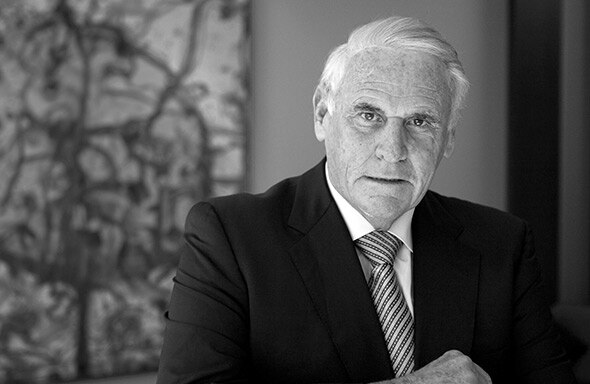 Lang Walker wears a suit and looks down the barrel of the camera. In the background is some kind of painting on the wall.