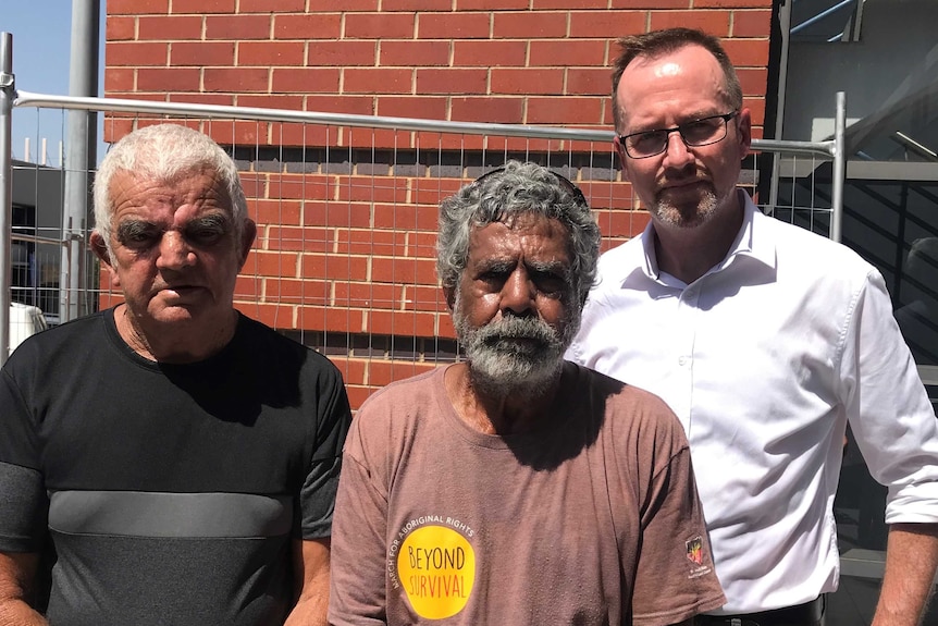 Three men with frowns stand in front of a brick wall on a sunny day.