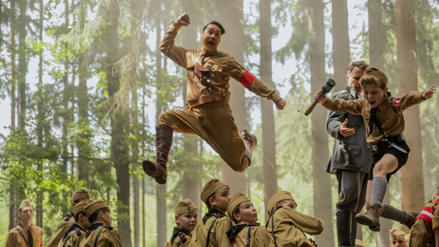 Forest scene with group of boys wearing Hitler Youth uniforms and Taika Waititi (as Hitler) and little boy leaping in air above.