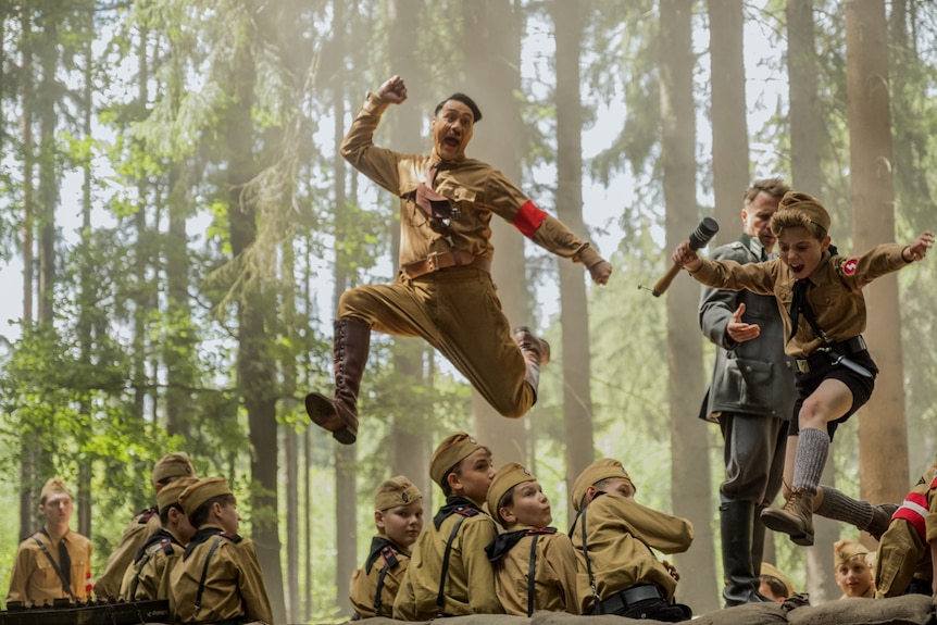 Forest scene with group of boys wearing Hitler Youth uniforms and Taika Waititi (as Hitler) and little boy leaping in air above.