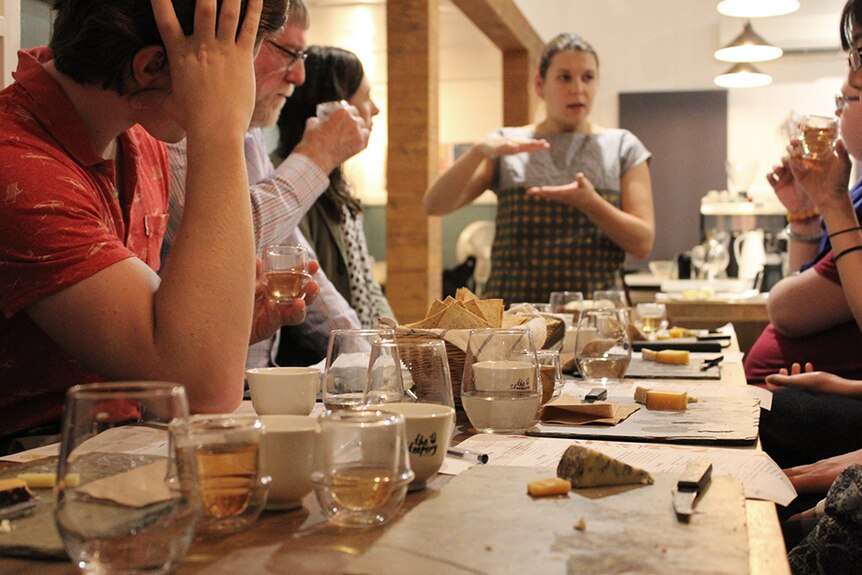 A group of people sitting around cheeseboards on a wooden table, eating and drinking.