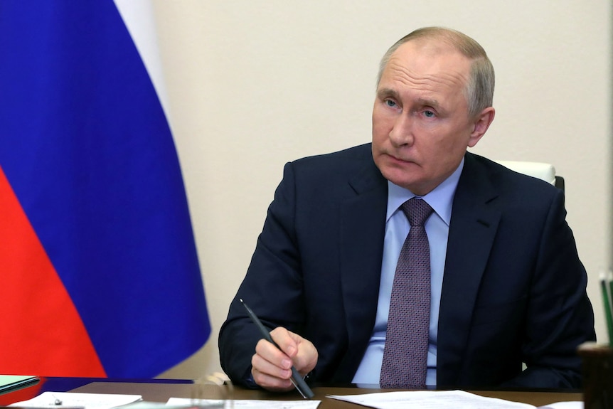 Sitting in front of a russian flag, Putin listens holding a pen