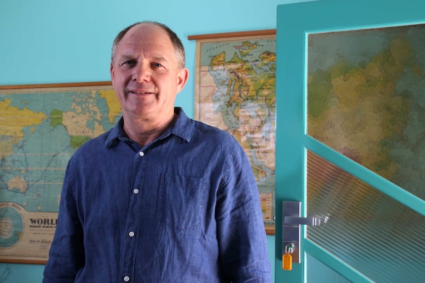 A mid-shot of David Sharp standing indoors in front of maps on the wall and next to a blue glass door.