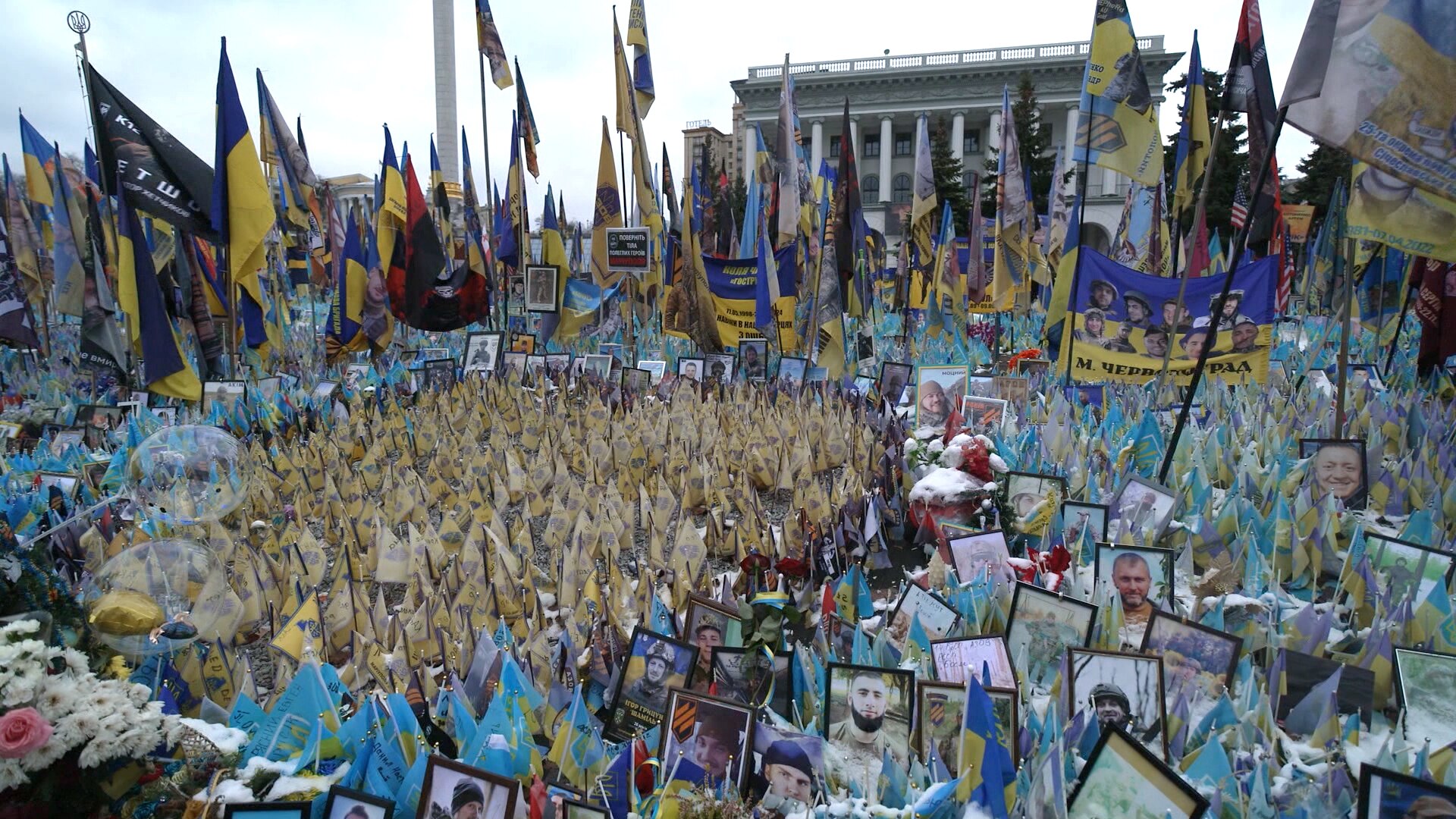 Hundreds of miniature flags and framed portraits spill out over a public square. Surrounding them are Ukrainian flags.