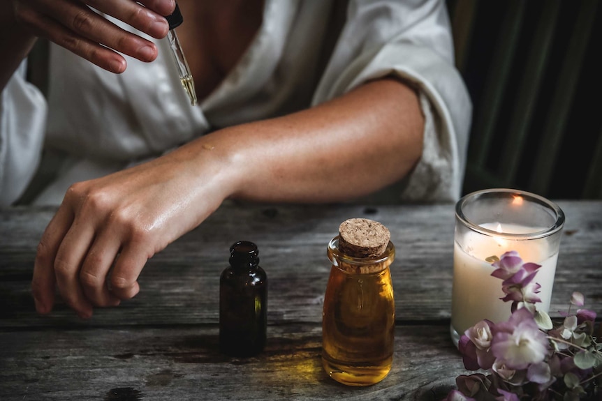 In a darkened room lit by a small candle, a woman uses an eye-dropper to put oil on her tanned skin.