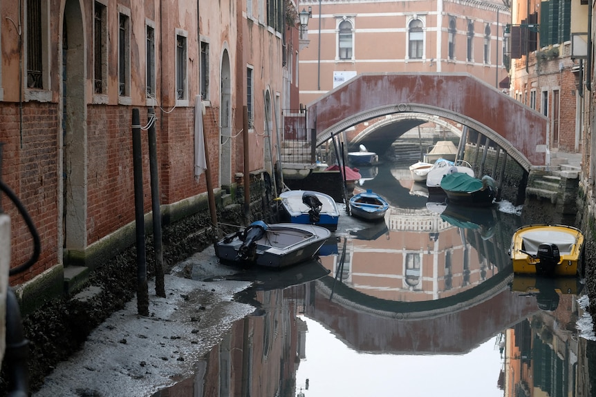 Varias pequeñas lanchas a motor y botes de remos se sientan dentro o al lado de una pequeña cantidad de agua en un canal veneciano.