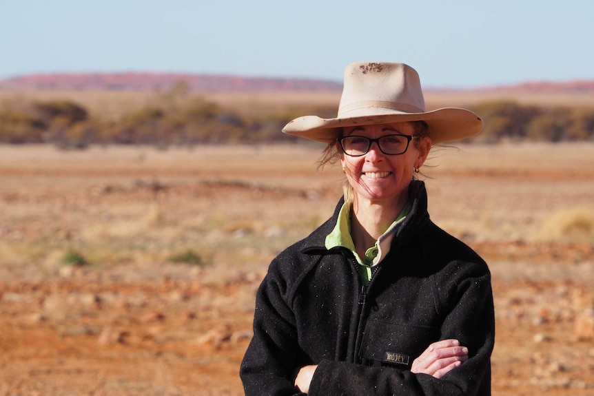 A lady in a broad hat stands in the middle of a brown and orange plain, wearing glasses and smiling wide.