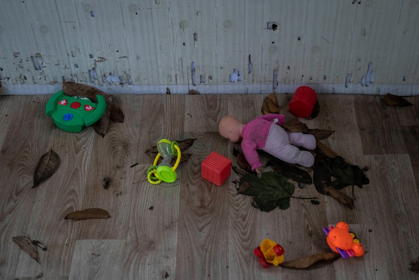 Toys, including a doll, lay on a wooden floor surrounded by leaves. 
