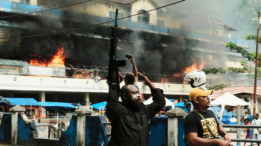 A man raises his rifles as a local market is seen burning in the background.