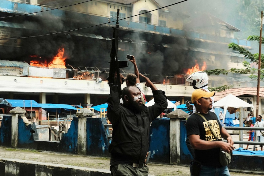 A man raises his rifles as a local market is seen burning in the background.