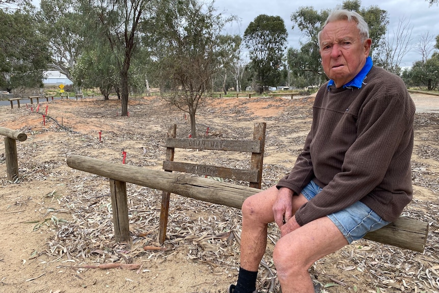 An older man sitting on a fence looking sad with a bare patch of land behind him.