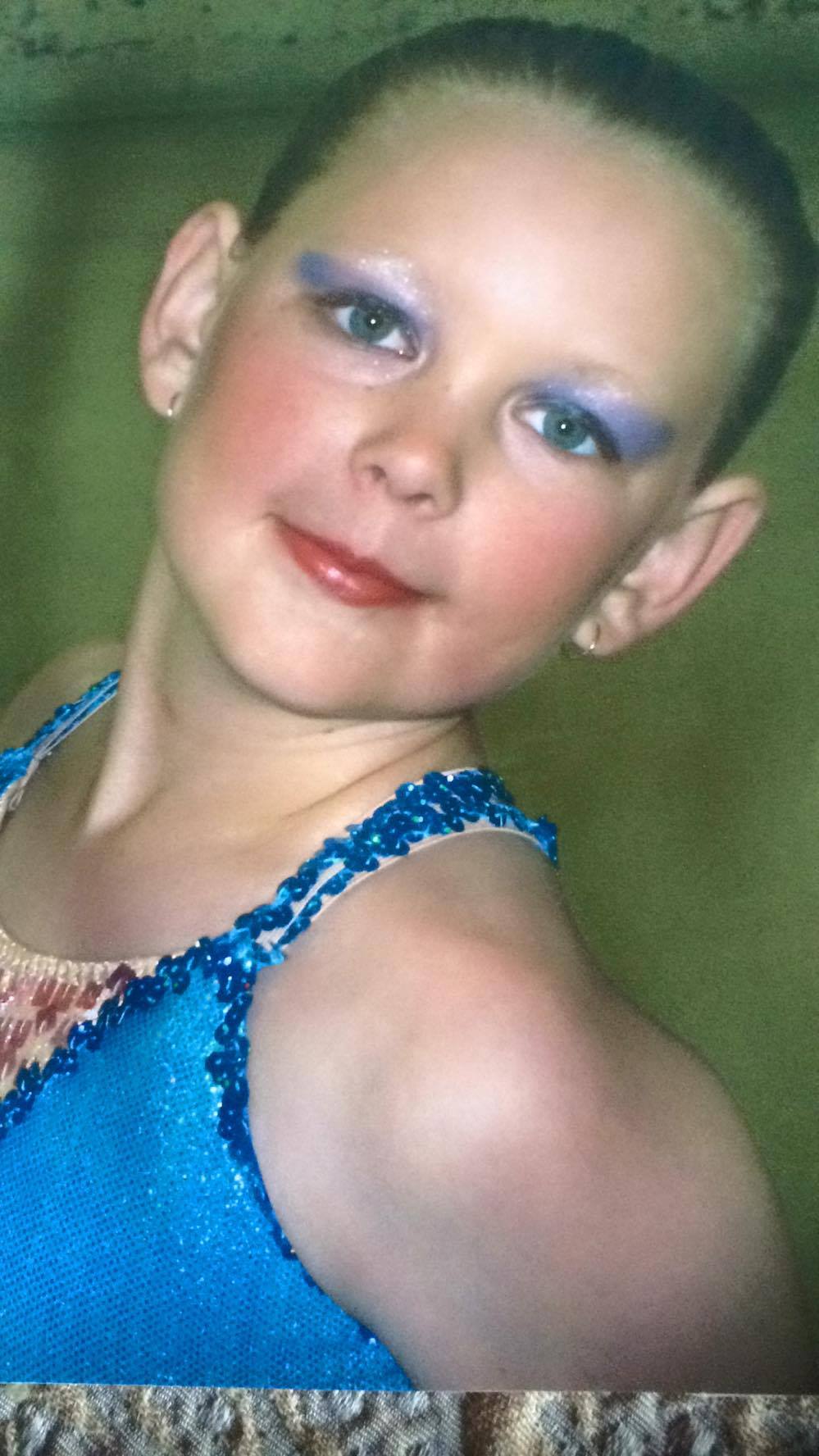 A headshot of a little girl smiling at the camera with over the top blue eye shadow and matching blue costume.