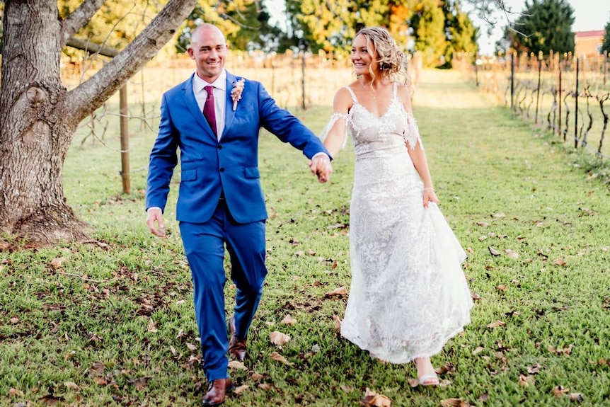A man dressed in a suit and a woman dressed in a bridal gown walk through a vineyard.