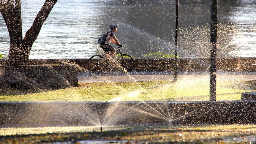 The morning sun shines through sprinklers as a cyclist rides along the promenade at South Bank.