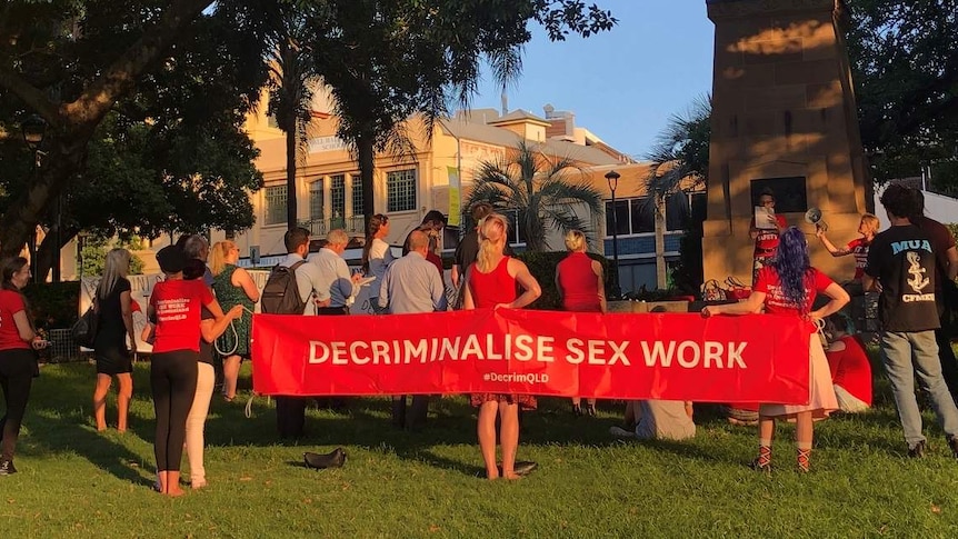 A group of people holding a sign, which reads "Decriminalise sex work" stand in a park.