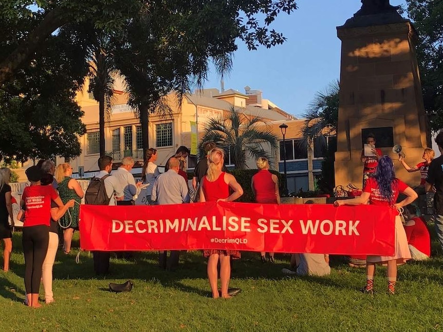 A group of people holding a sign, which reads "Decriminalise sex work" stand in a park.